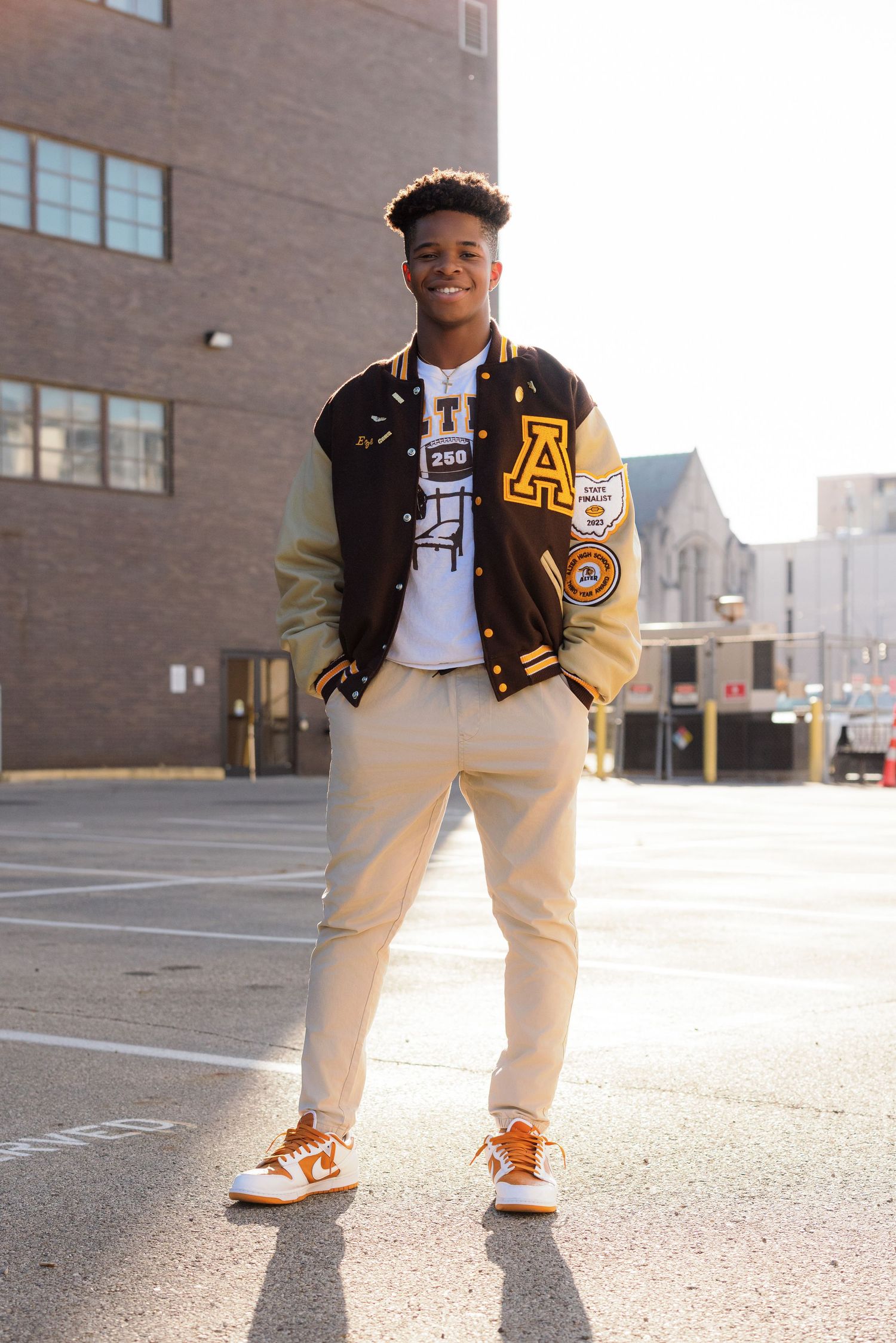 A person wearing a varsity letterman jacket and khaki pants stands confidently in an urban setting.