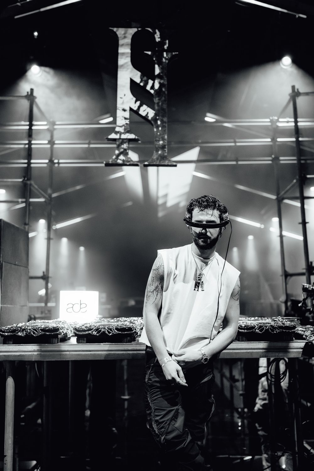 DJ performing behind decks in white outfit with dramatic stage lighting creating moody atmosphere.