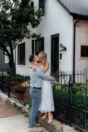 A couple embraces by a white building with black shutters and iron fencing.