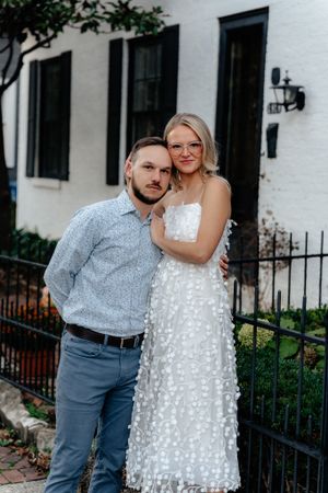 A couple poses together against a white building with black shutters.