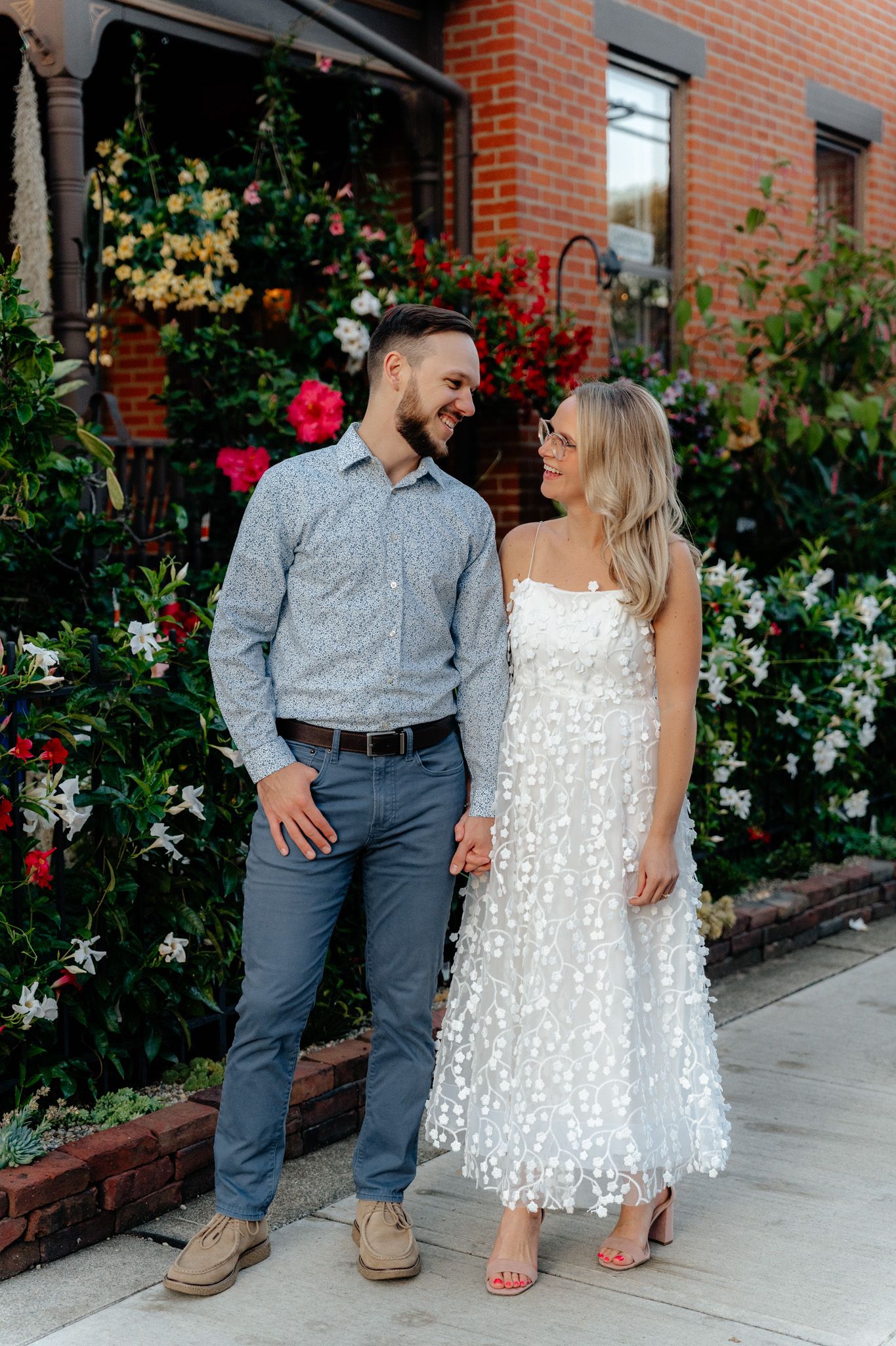 A couple stands close together on a sidewalk with flowering bushes behind them.