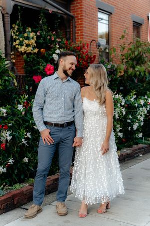 A couple smiles at each other in front of a brick building with colorful flowering garden.