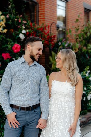 A couple shares a romantic moment in front of a red brick building with colorful flowering bushes.