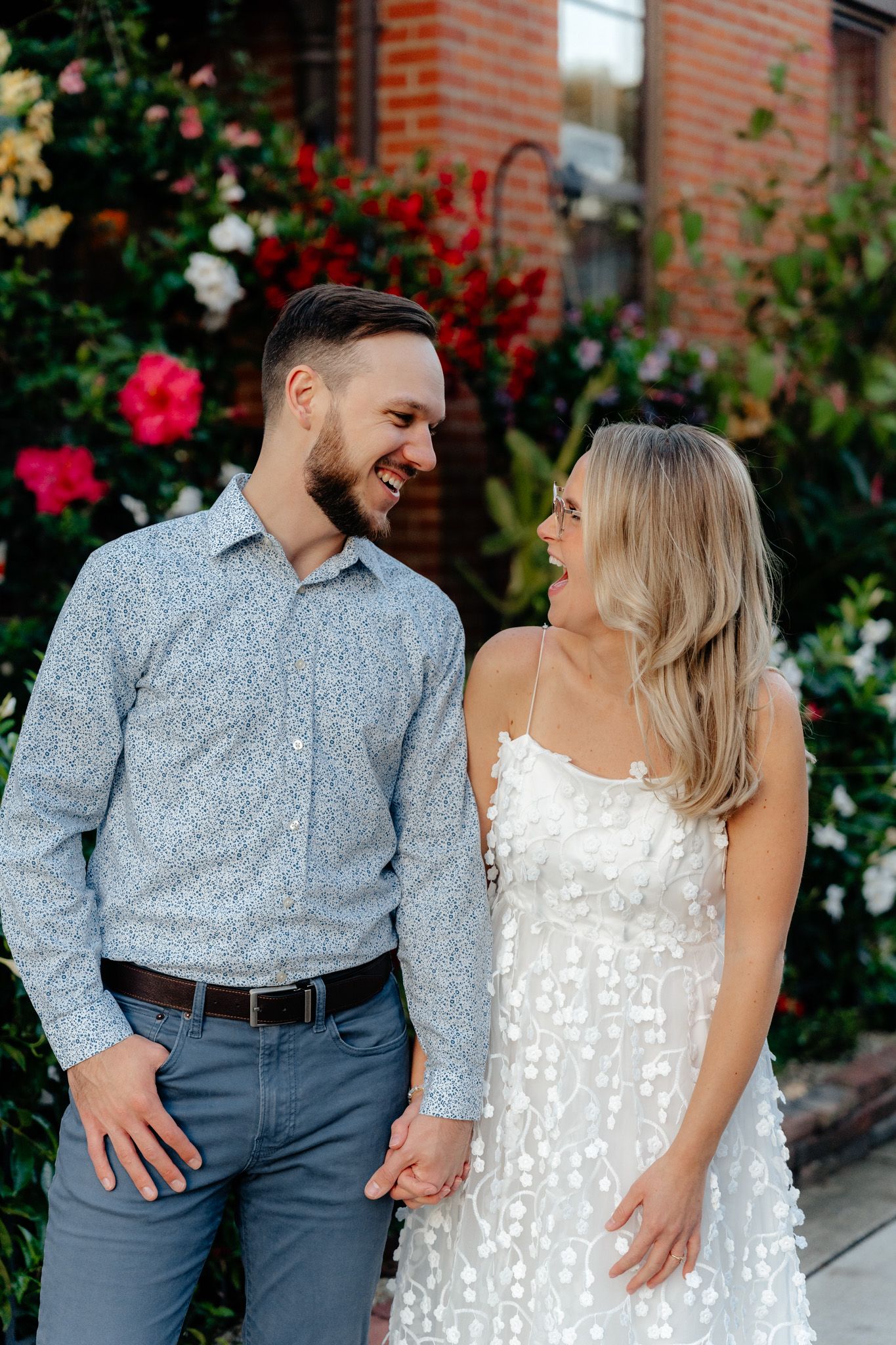 A couple gazes lovingly at each other while standing beside vibrant floral landscaping.