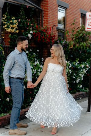 A couple laughs together while standing in front of a blooming garden against brick architecture.