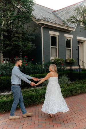 A romantic dance pose on a brick sidewalk in front of a charming grey house with a white lace dress and casual attire.