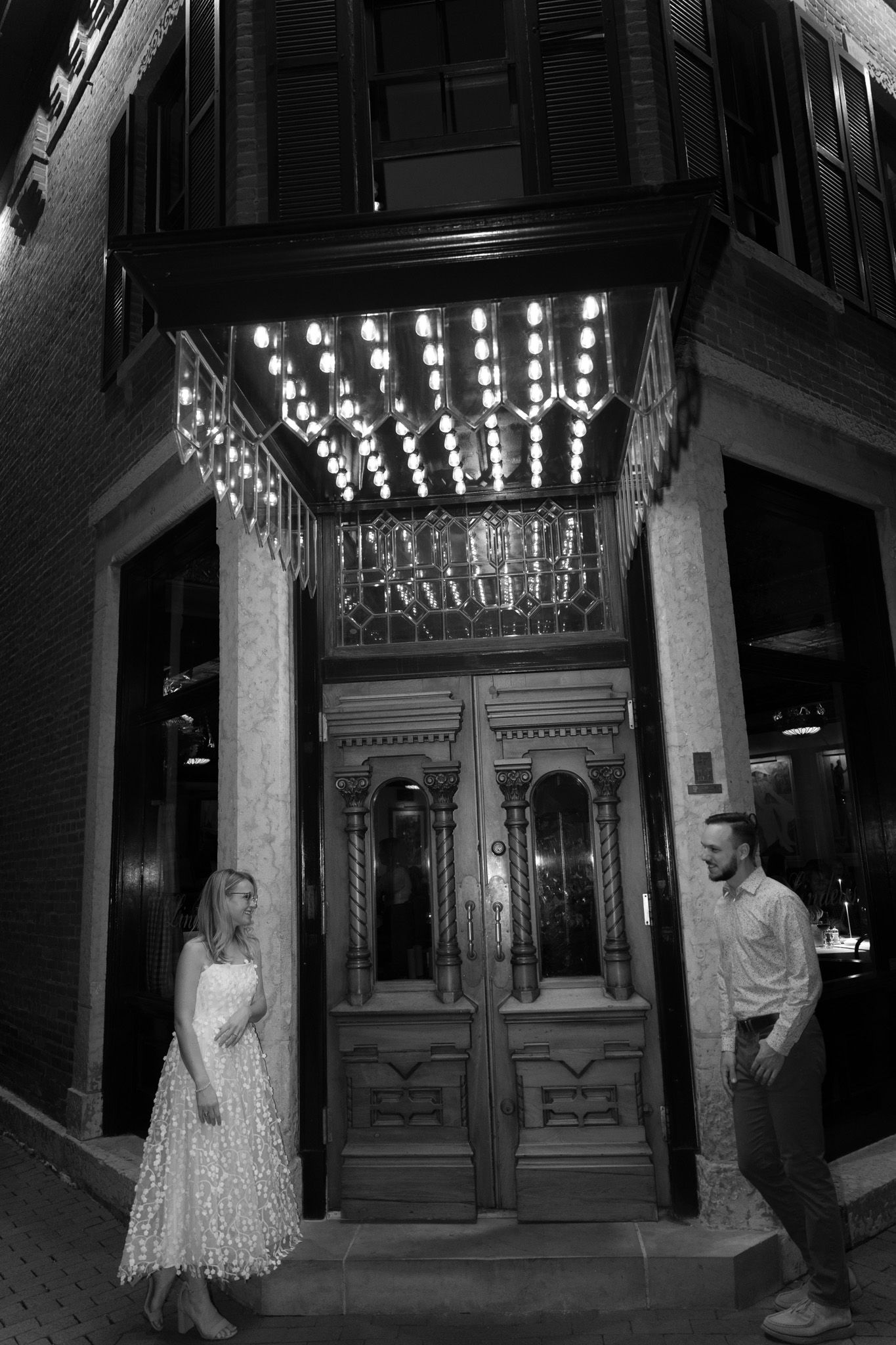 A dramatic black and white portrait under an ornate lit entrance with vintage doors.