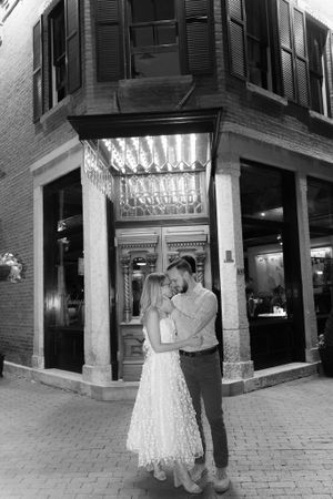 Couple embraces romantically outside an illuminated storefront window at night on a brick sidewalk.