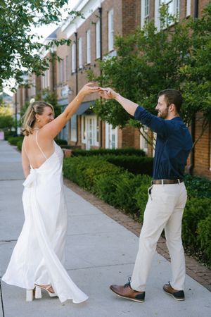 A couple in formal attire dances playfully on a sidewalk in front of brick townhouses.