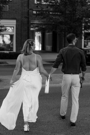 A couple walking away from camera holding hands, with her white dress flowing in the breeze.