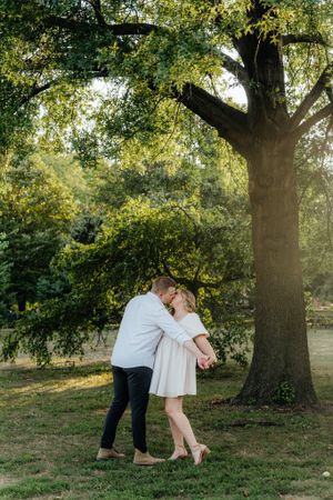 A romantic couple embraces under a large tree in a sunlit park setting.