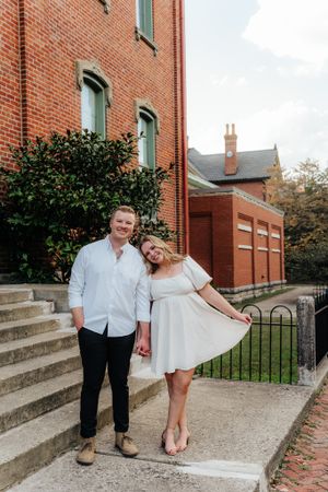 A couple poses playfully on stone steps in front of a red brick building.