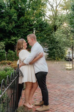 A romantic couple embraces near an iron fence on a brick pathway surrounded by lush greenery.