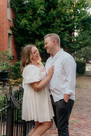 A couple shares a lighthearted moment beside an iron fence near a brick building.