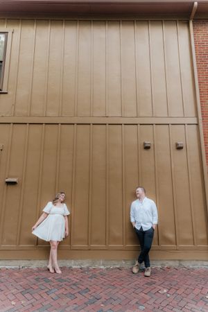 A couple poses playfully against a large tan wall on a brick pathway.