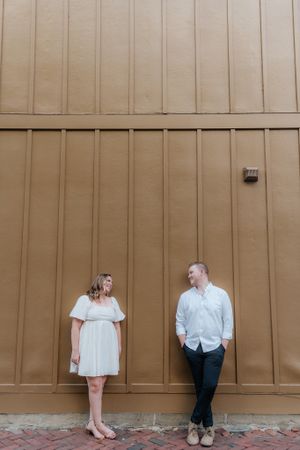 A couple stands separated against a tall tan wall on brick pavement.