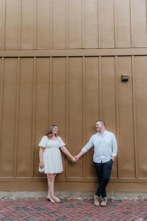 A couple holds hands while standing against a large tan wall outdoors.