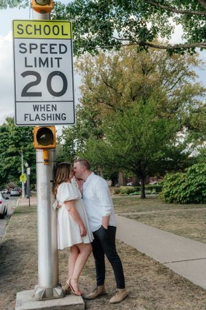 A couple shares a romantic moment by a school speed limit sign surrounded by trees.