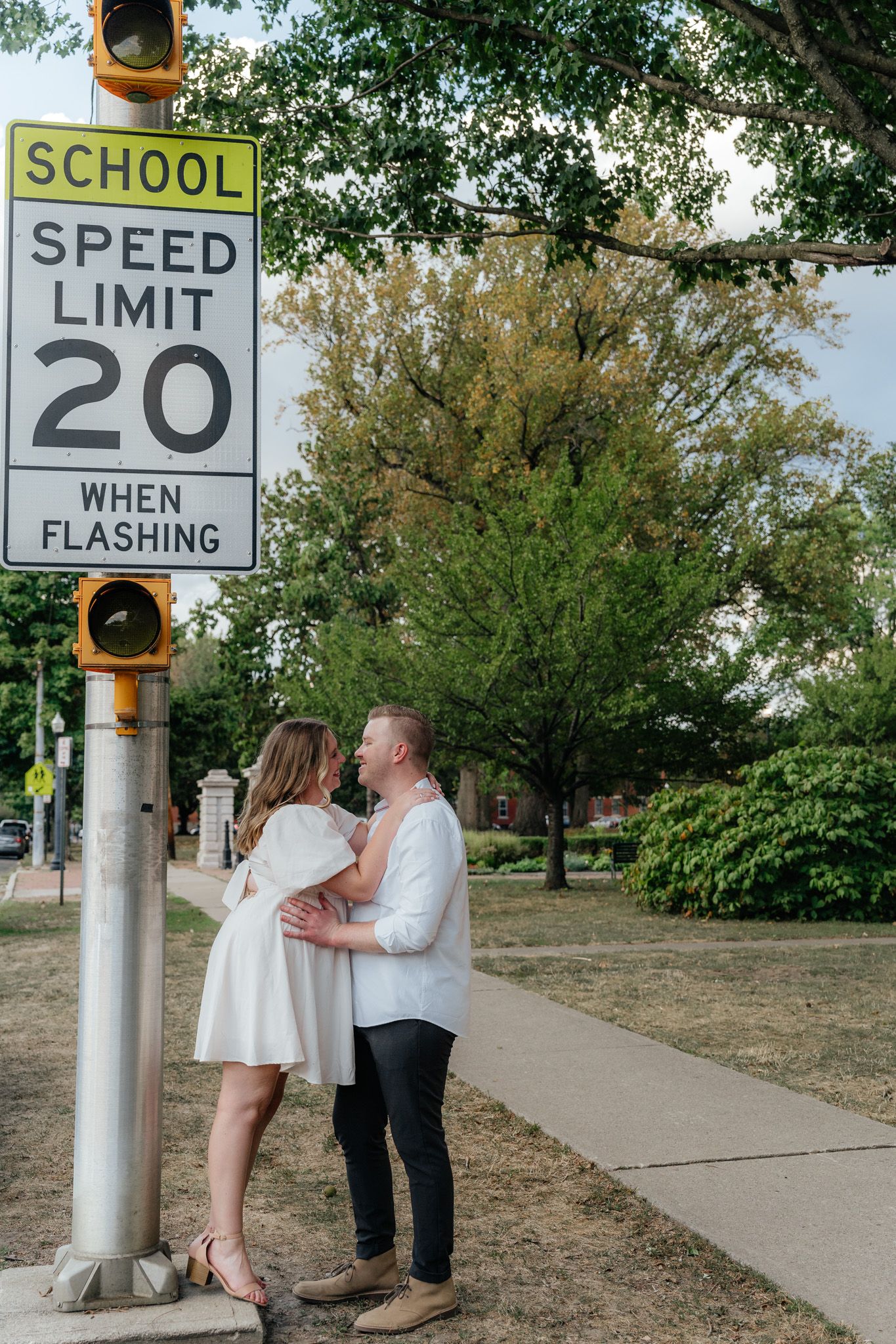 A couple embraces near a school zone sign on a tree-lined sidewalk.