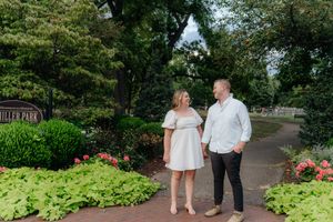 Engaged couple strolling along garden path with blooming flowers and greenery.