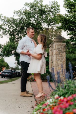 A romantic couple in light summer attire pose together near a stone pillar and colorful flowers.