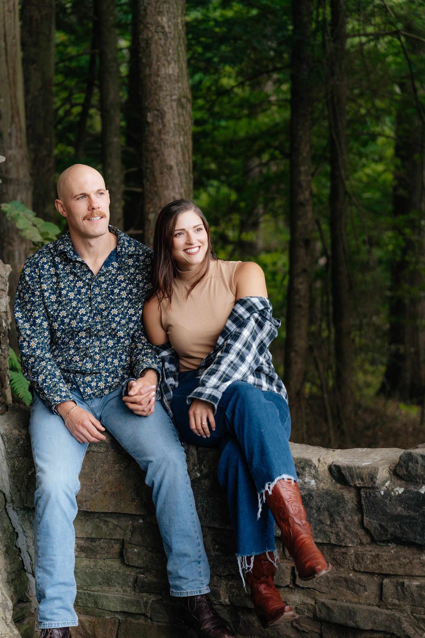 A couple sitting together on a stone wall in a wooded setting wearing casual fall attire.