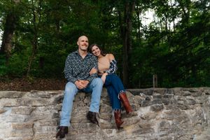 A relaxed outdoor portrait on a stone wall surrounded by lush green trees.