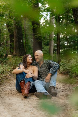 A couple sits together laughing on a forest trail surrounded by lush green trees.