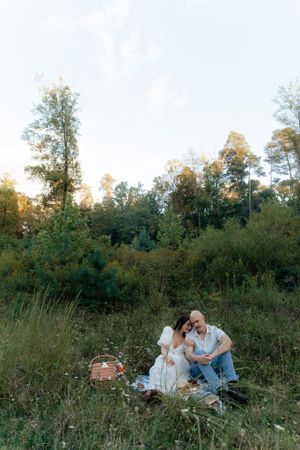 A couple enjoys a sunset picnic in a natural meadow setting with tall trees.