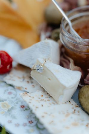 Close-up of an engagement ring placed on brie cheese with fruit and jam on a picnic spread.