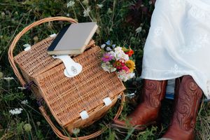 Wicker picnic basket with flowers and brown leather cowboy boots on grass.