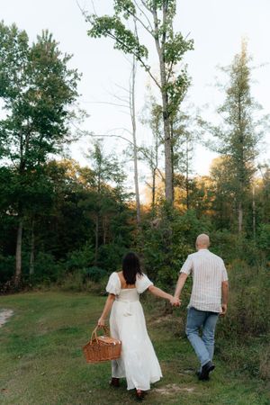 A couple walking hand in hand through a wooded path carrying a picnic basket at sunset.