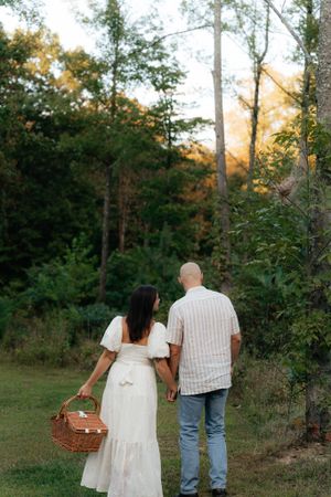 A couple stands together in a forest clearing carrying a wicker picnic basket.