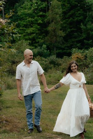 A couple holds hands while walking through a lush green forest setting.