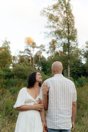 A couple embraces while standing in a meadow surrounded by trees during golden hour.