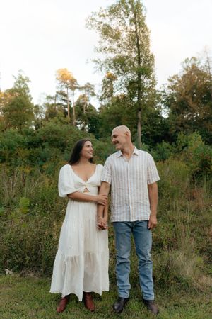 A couple poses together in front of a wooded area wearing contrasting white and striped outfits.