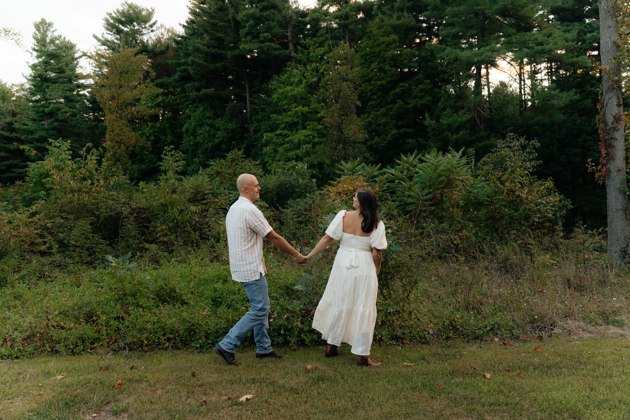 A couple walks hand in hand through a grassy field with evergreen trees in the background.