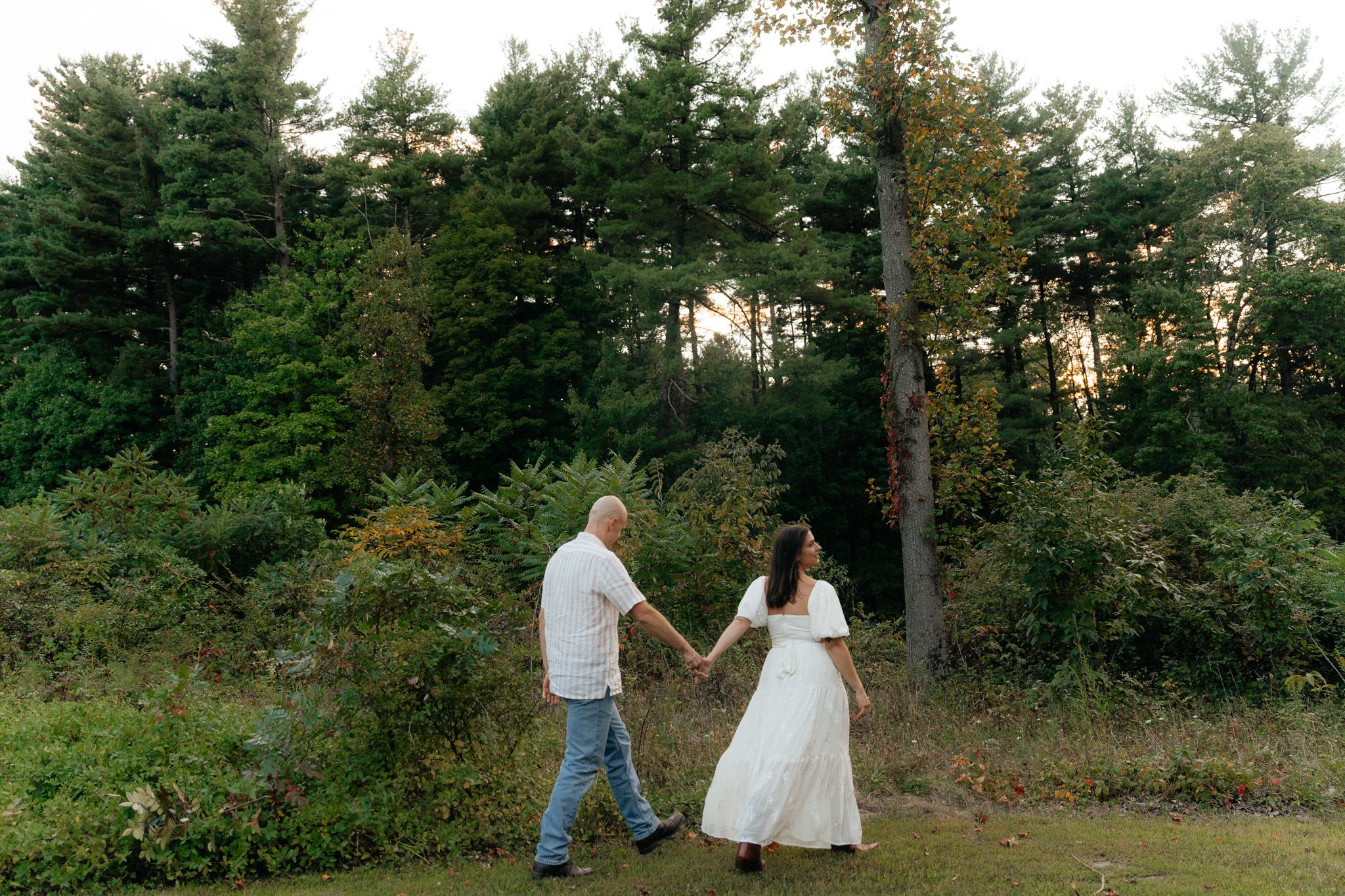 A romantic couple walks hand in hand through a pine forest at sunset in casual and formal attire.
