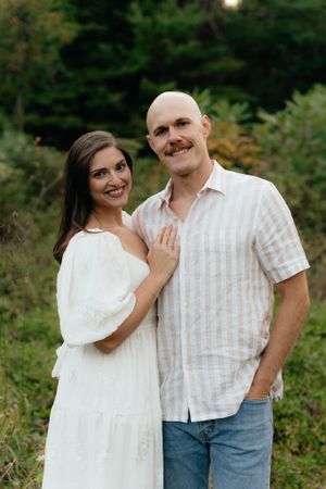 A couple poses for a portrait in front of lush green forest wearing coordinated white and beige outfits.