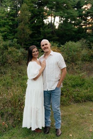 A loving couple stands together in a forest clearing during golden hour.
