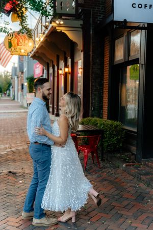 Couple embraces on brick sidewalk outside vintage storefront at sunset.