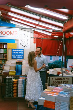 Romantic moment in bookstore aisle surrounded by stacks of books and fluorescent lighting.