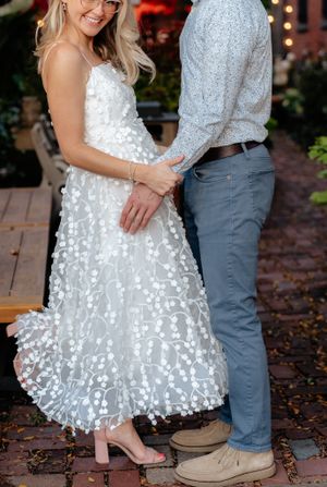Close-up detail of a textured white wedding dress with floral appliques next to casual grey pants.