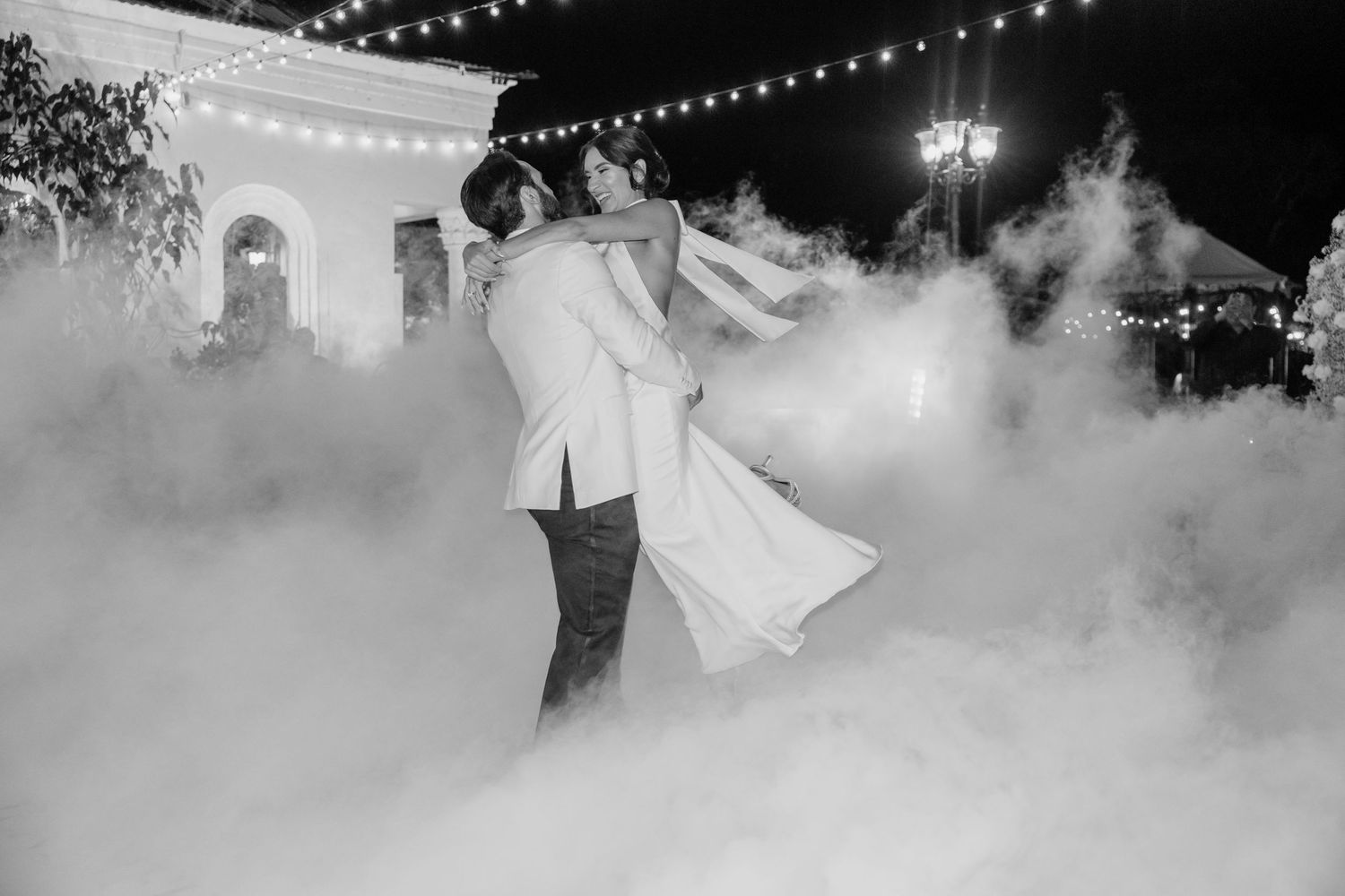 A couple dances their first dance on a misty dance floor illuminated by string lights at an outdoor wedding reception.