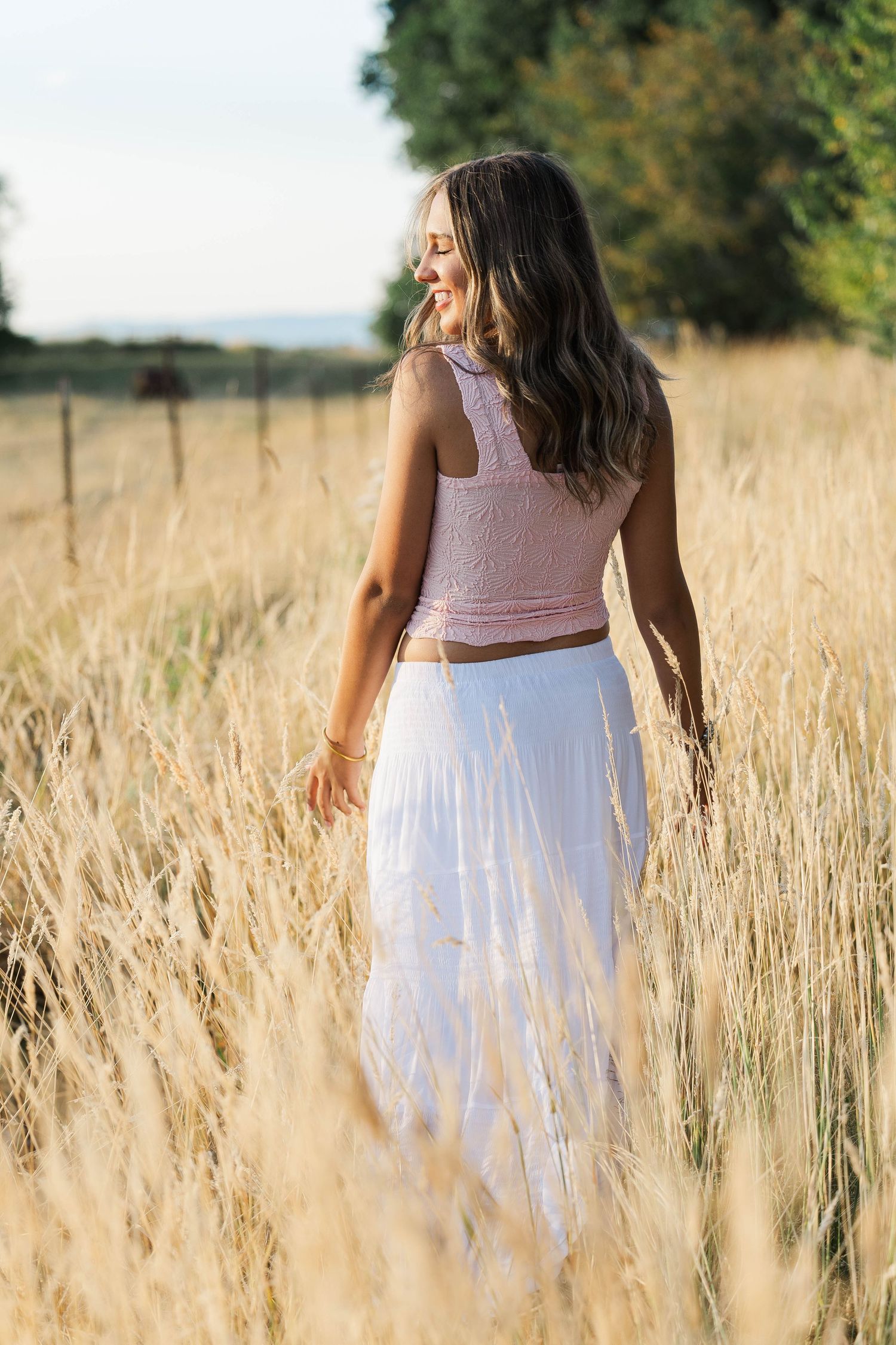 Young woman in pink crop top and white maxi skirt walks through tall golden grass in a sunny field.
