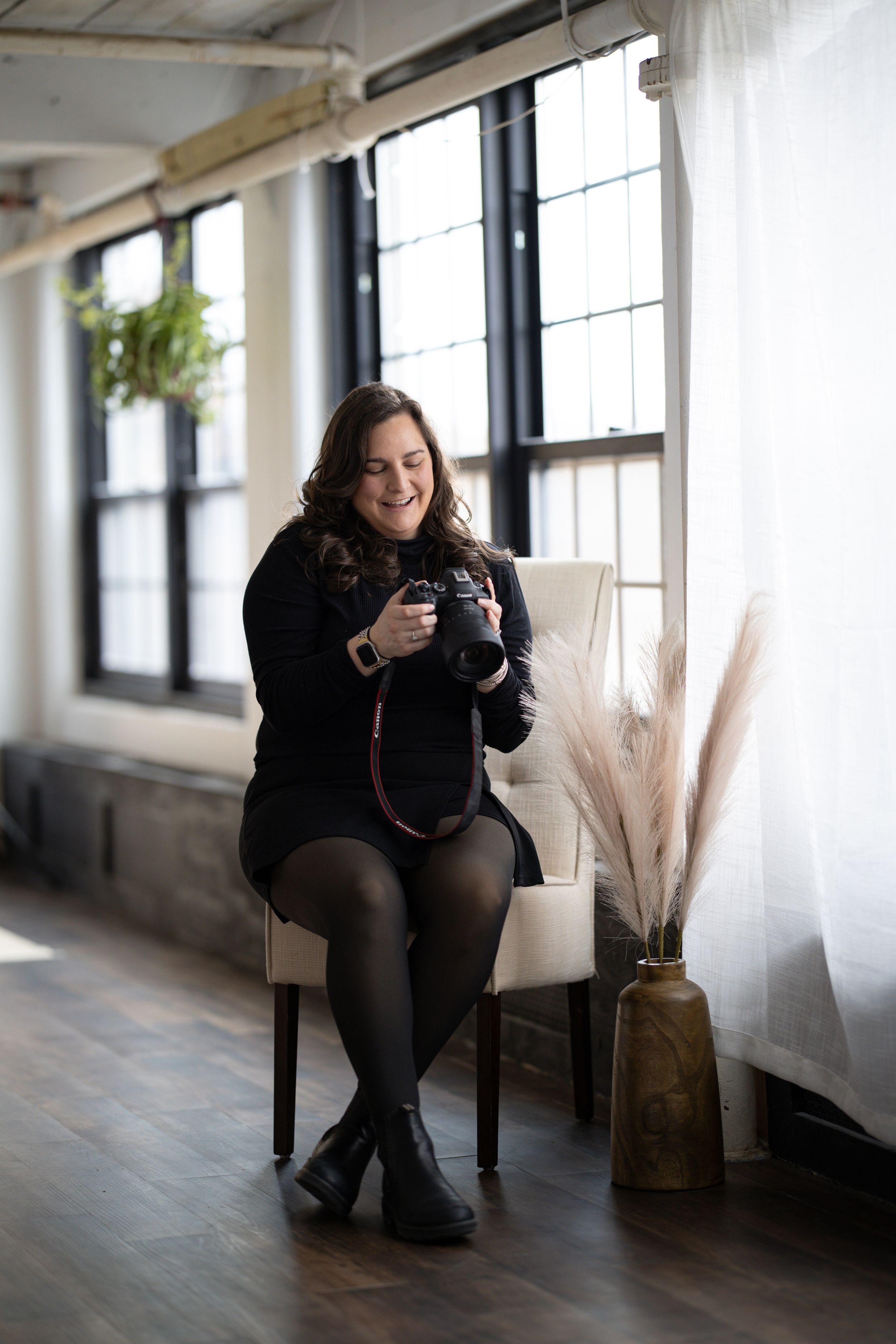 Smiling female photographer sitting in chair holding camera in bright studio with large windows.