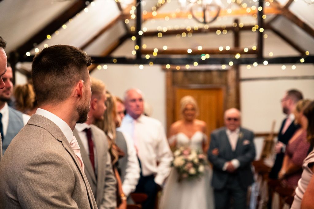 Bride walking down the aisle at Coton House Farm by an older man, with guests looking on a rustic, fairy-light decoratio
