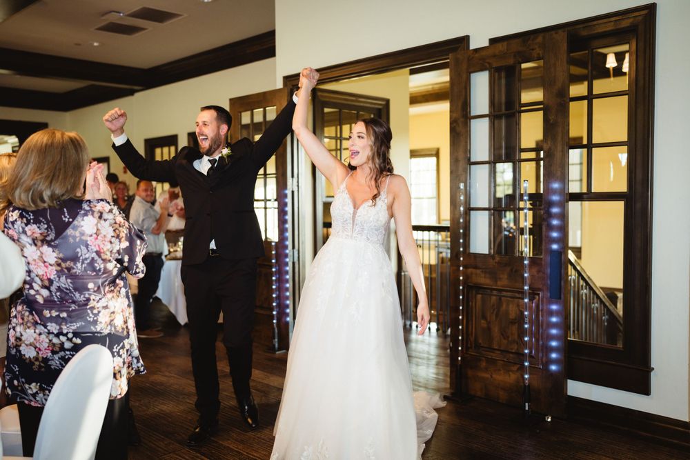 Newlyweds make a joyous entrance at their wedding reception with raised arms while guests cheer.