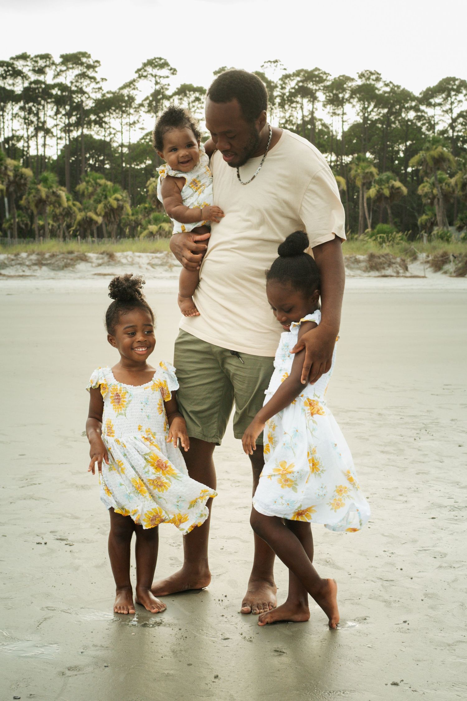 A family enjoys a tender moment together on a sandy beach with pine trees in the background.