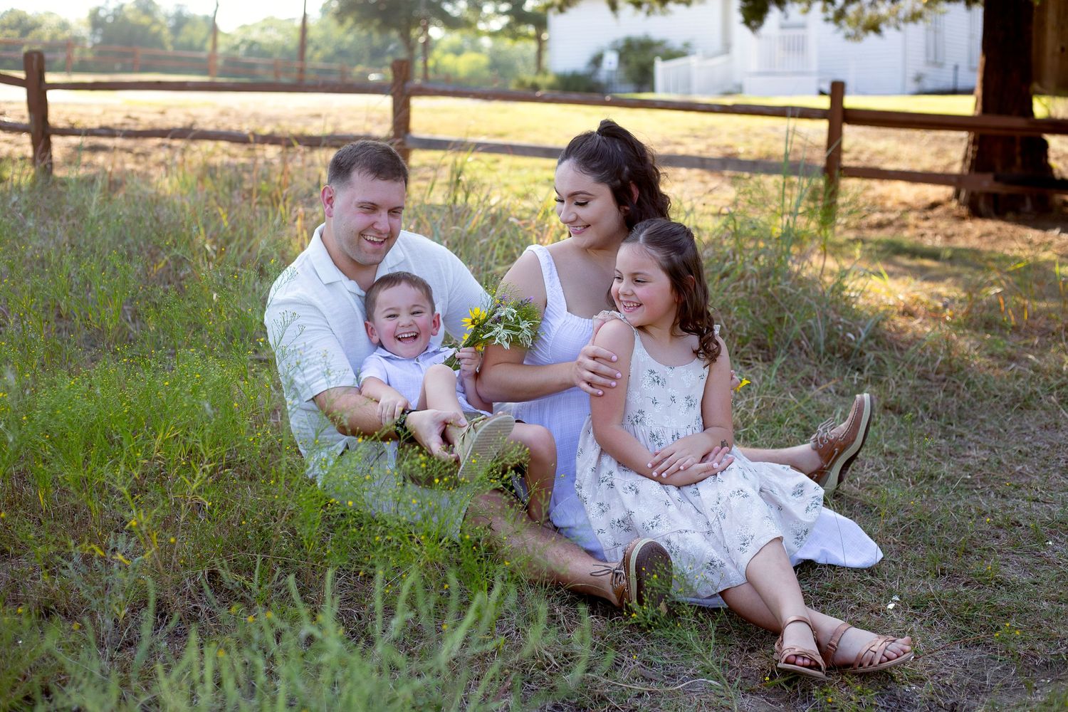 A family enjoys a relaxing moment together sitting in tall grass near a wooden fence at sunset.